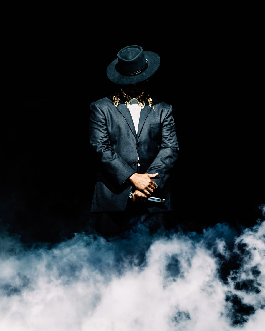 Ne-Yo standing still in a grey suit and wide-brimmed hat, head lowered, low fog rolling across the stage in front of him