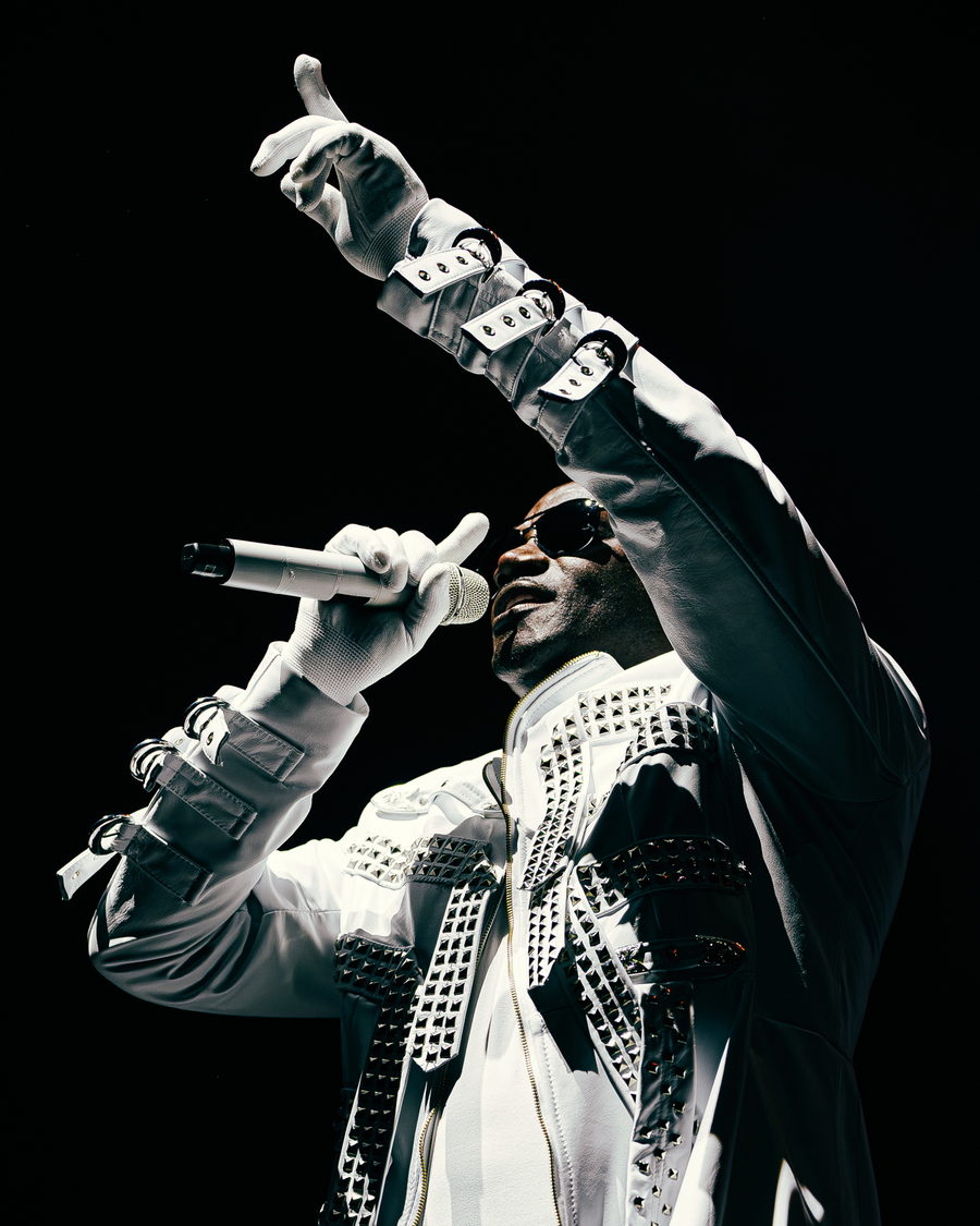 Akon shot from below with one gloved arm raised into the light, his white studded coat blazing against a black stage