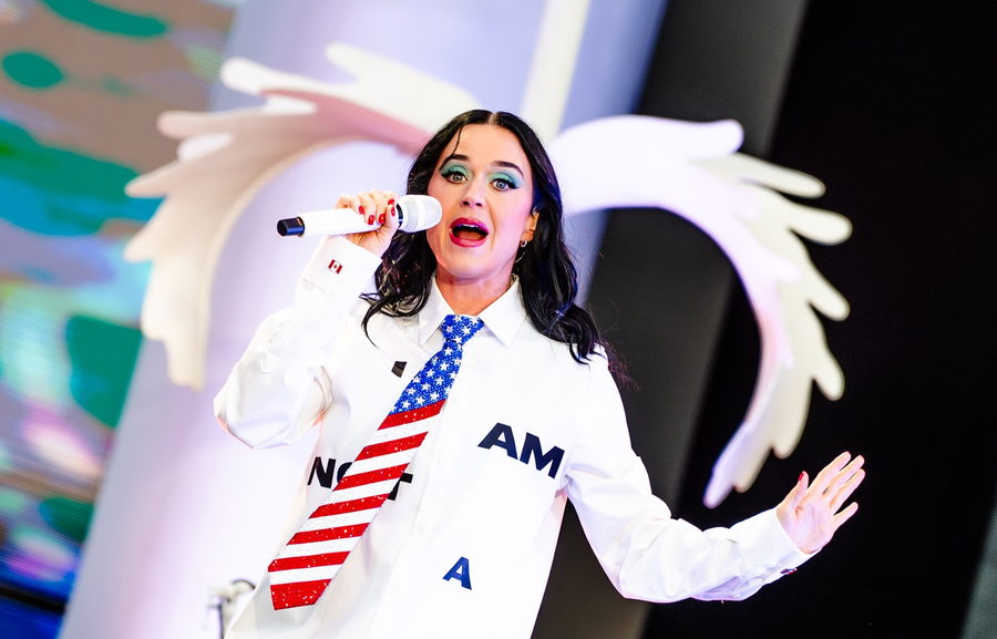 Katy Perry mid-line with a hand raised, a large white sculpted wing curving around her on the stage set