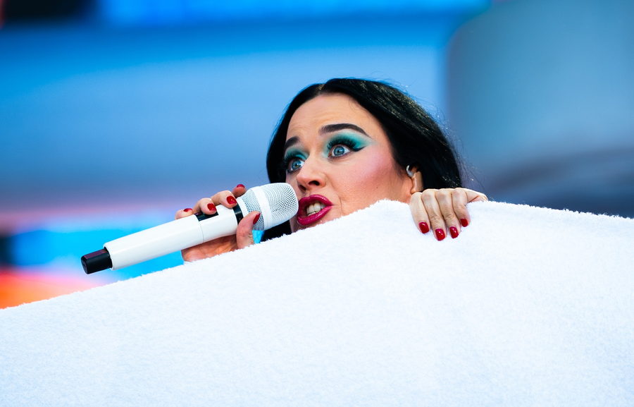 Katy Perry peering over a white prop cloud with a microphone at her lips, green eye make-up and red lipstick