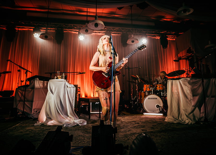 Hayley Williams at the microphone with a cherry-red guitar, the stage dressed with draped white sheets and orange curtains, the drummer behind her