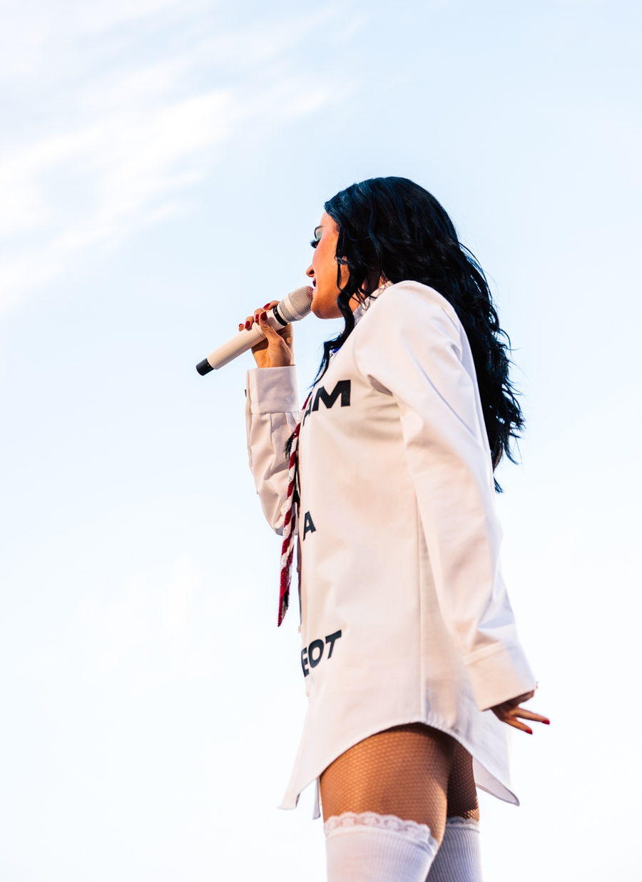 Katy Perry in profile against a pale open sky, black hair blown back, singing into a white microphone