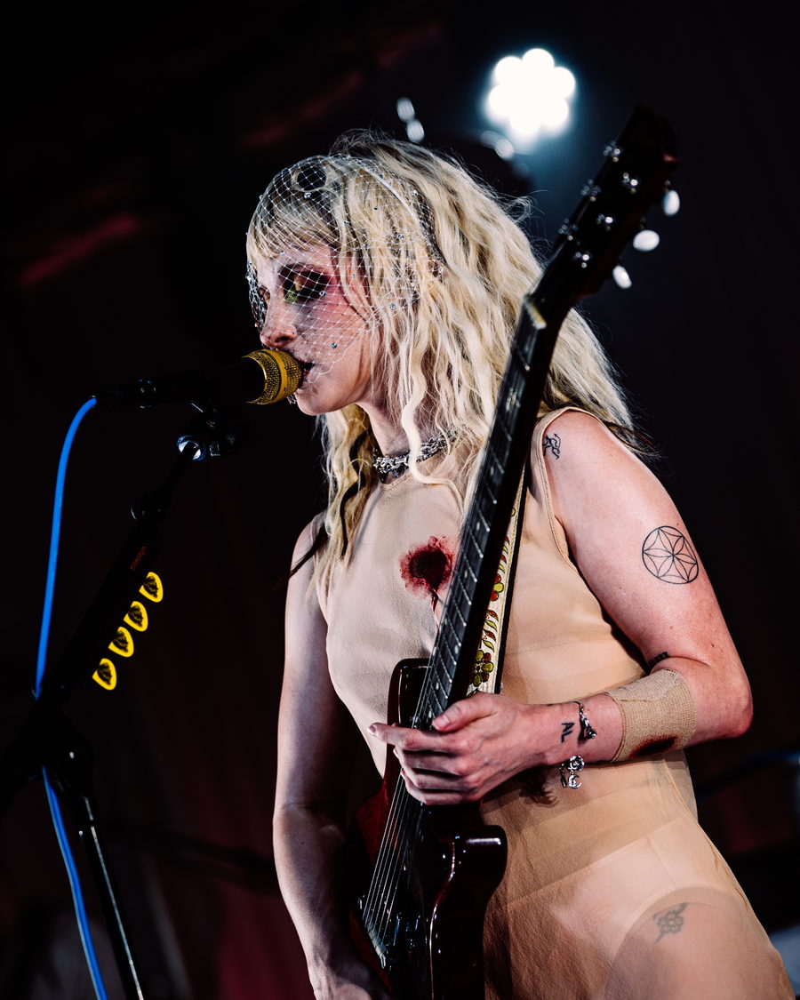Hayley Williams close up at a gold microphone, blonde hair loose under a jewelled veil, one hand on the neck of her guitar
