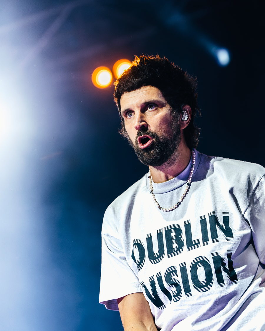 Close portrait of Kasabian's singer looking up, wearing a white t-shirt reading Dublin Vision, warm lamps and blue haze behind him