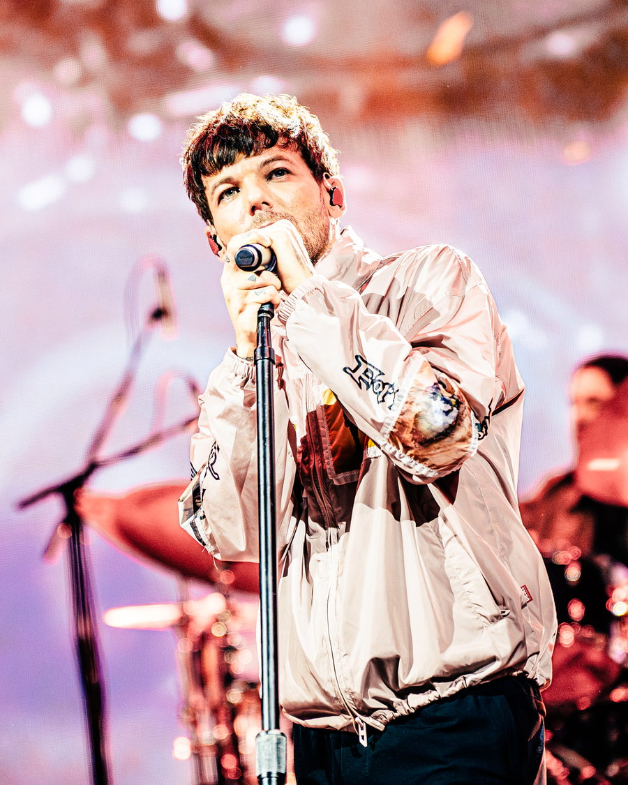 Louis Tomlinson close up at the microphone stand in a pink jacket, warm pink stage light and the drum kit blurred behind him
