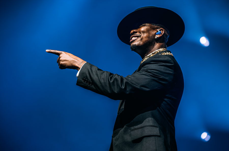 Ne-Yo in profile, smiling, pointing out into the room against a deep blue backdrop