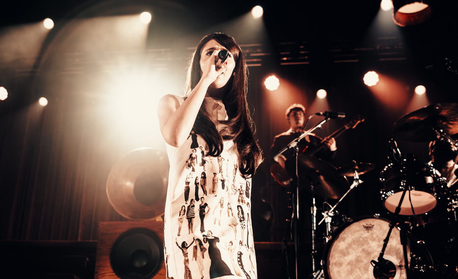 Sienna Spiro singing with the microphone raised to her face, a hard backlight flaring beside her and the drum kit behind