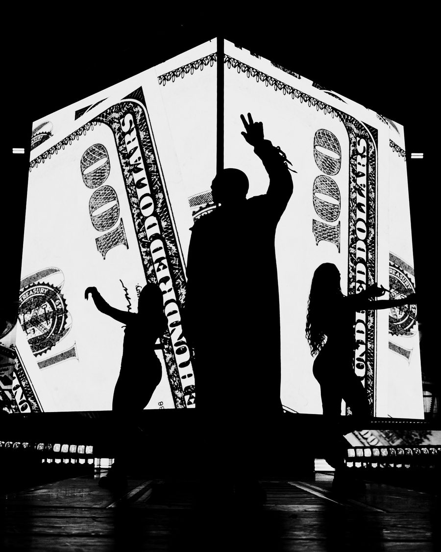Akon and two dancers in hard silhouette against screens filled with a hundred dollar bill, one arm raised