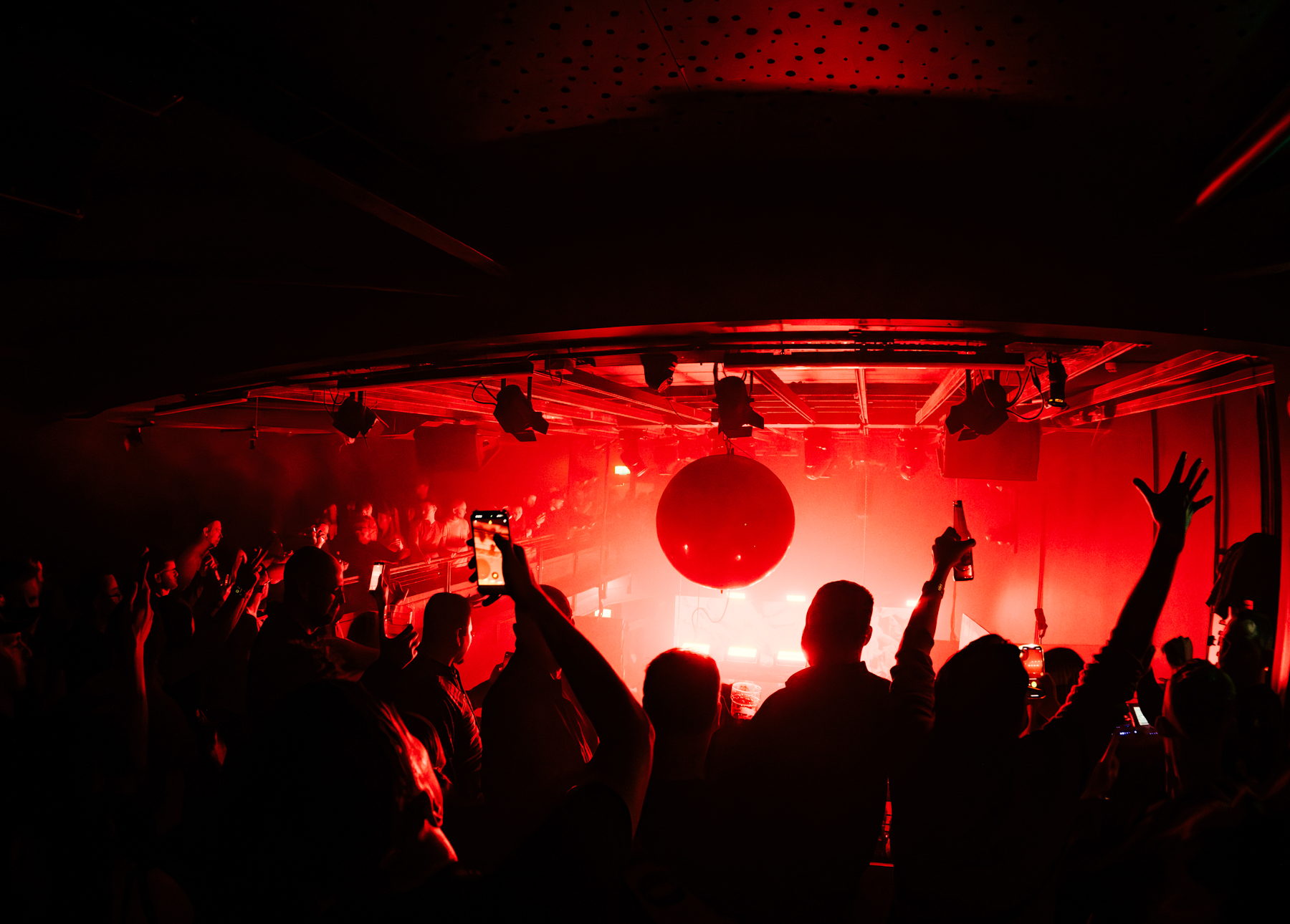 Club crowd silhouetted in deep red light with hands and phones raised towards a mirrorball
