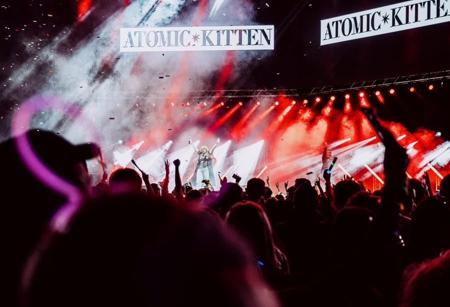Atomic Kitten on a red-lit stage seen over a festival crowd, the band's name on banners either side