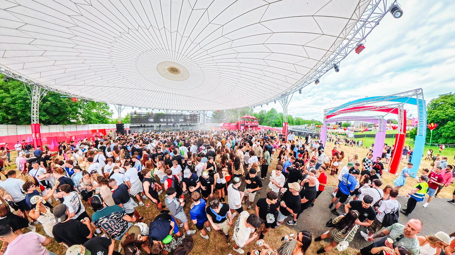 Fisheye view of a packed festival crowd under a wide white canopy, the main stage beyond it in the afternoon sun
