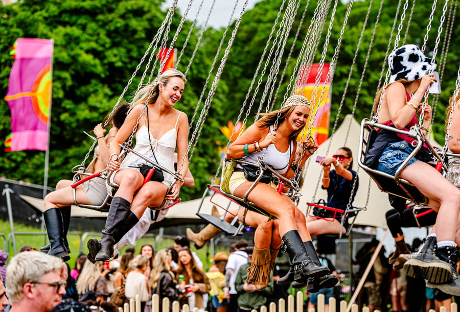 Two friends laughing side by side on a swing ride above the festival field, flags and tents behind them