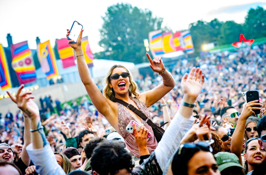 Someone lifted on shoulders above a huge daytime festival crowd, arms up and a phone in hand, painted banners on the stage behind