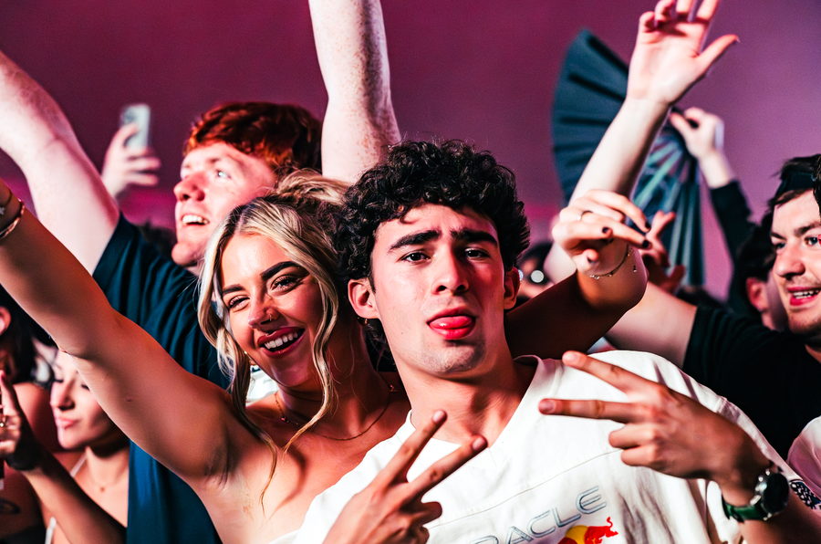 Two friends throwing peace signs in a tightly packed tent crowd washed in pink light