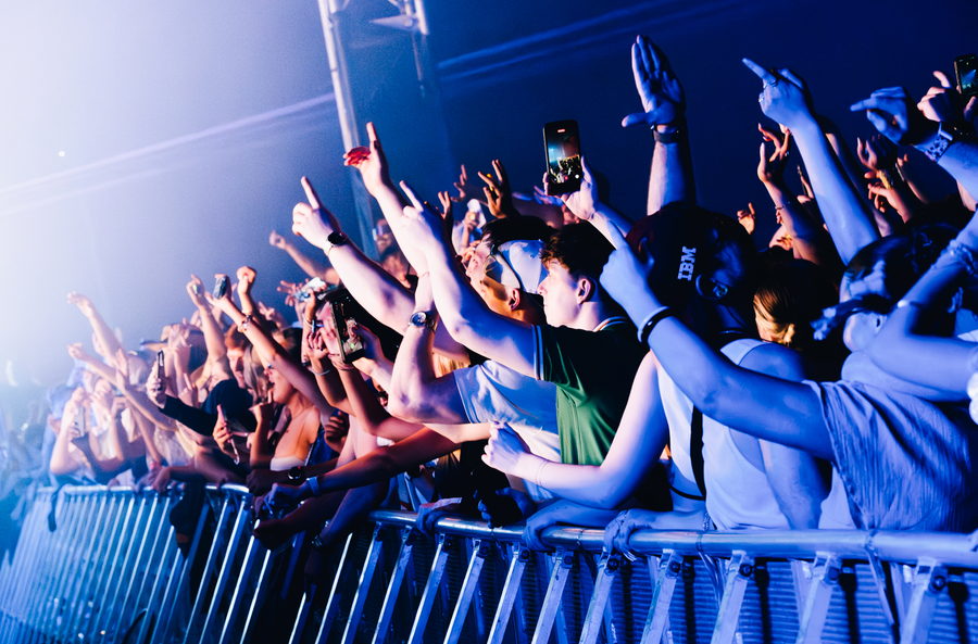 A line of fans along the front barrier with arms and phones raised, lit hard blue from the side of the stage