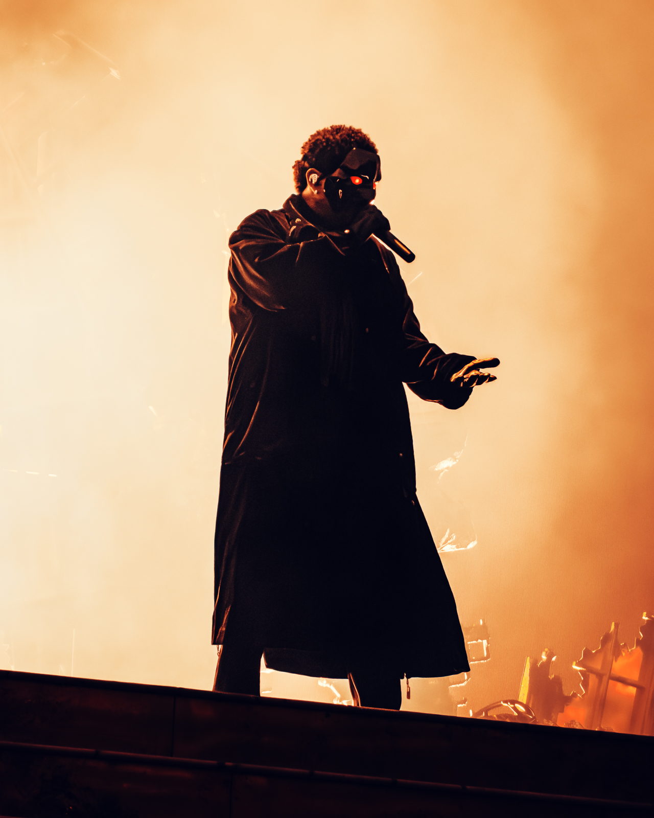 The Weeknd alone at the top of the stage in a long dark coat, lit from behind in amber haze