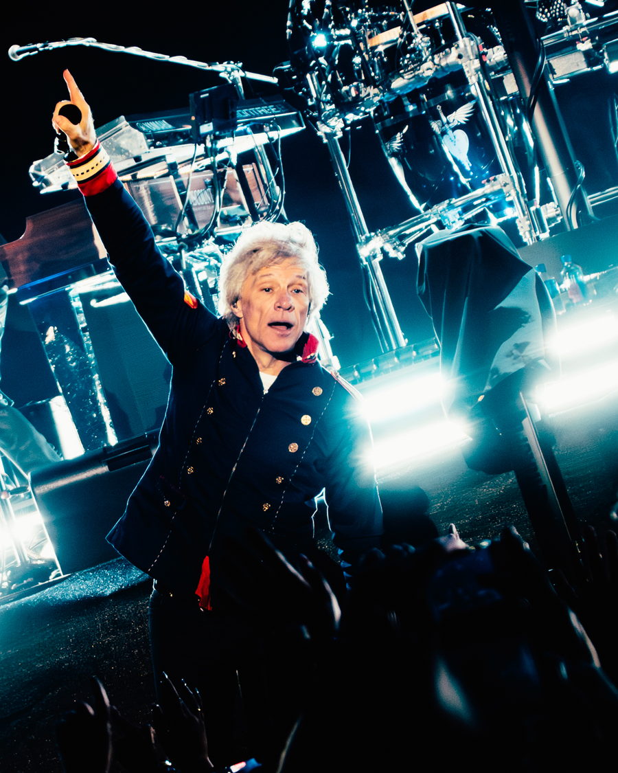 Jon Bon Jovi at the microphone with one arm raised, banks of white light behind him
