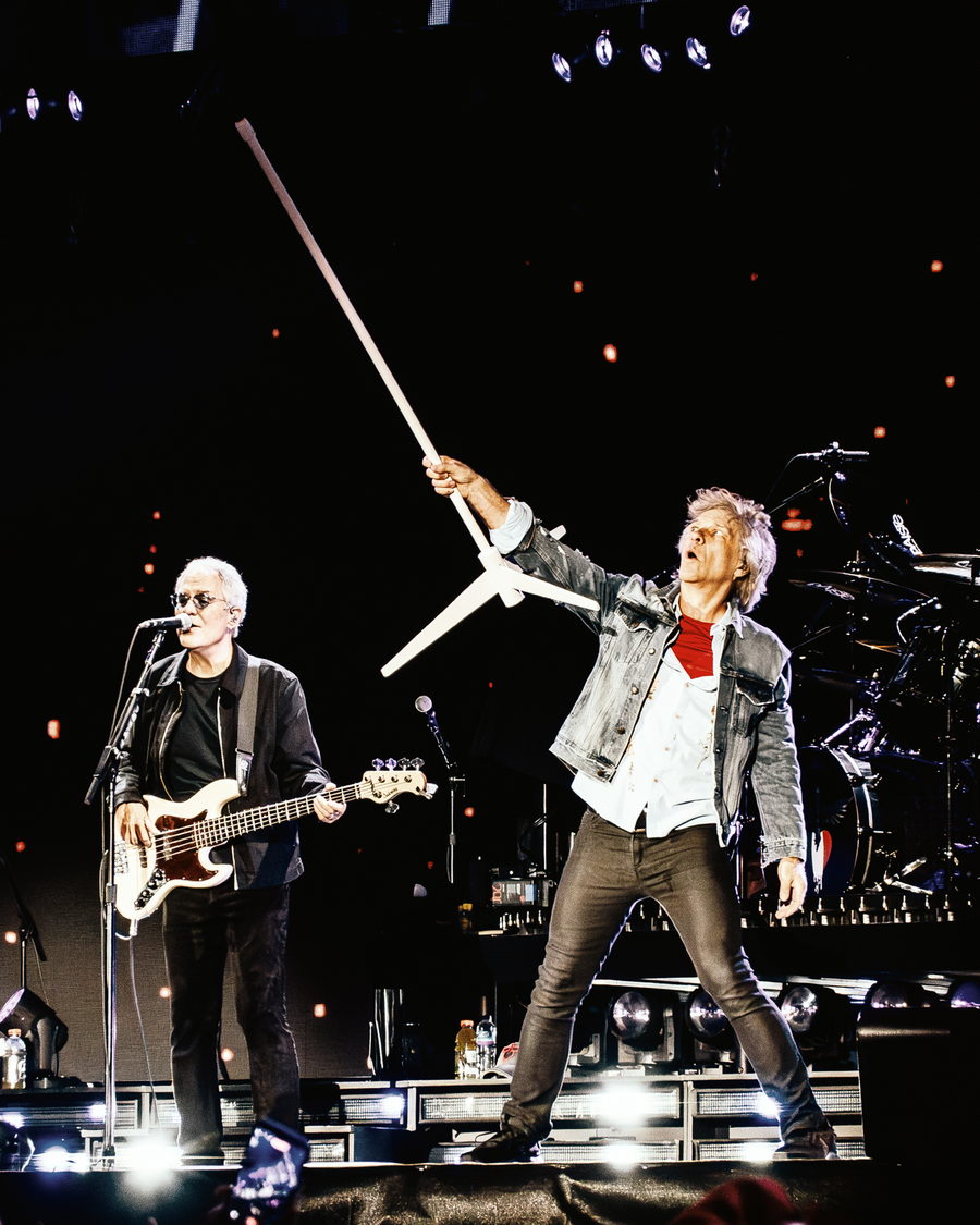 Jon Bon Jovi raising a hand beside his guitarist on a darkened arena stage