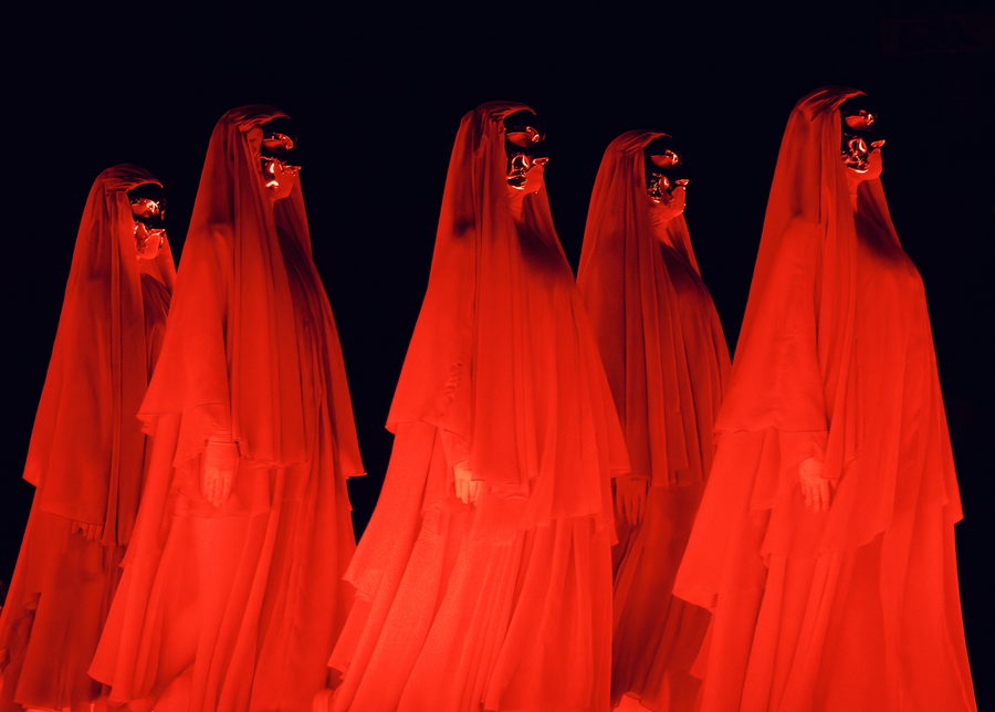 Rows of performers in red hooded robes lit from the side, filling the frame