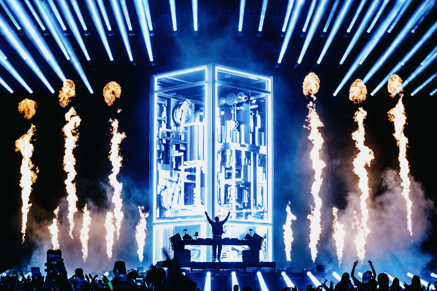 Flame jets rising either side of a lit DJ booth, blue strobes overhead and an arena crowd below