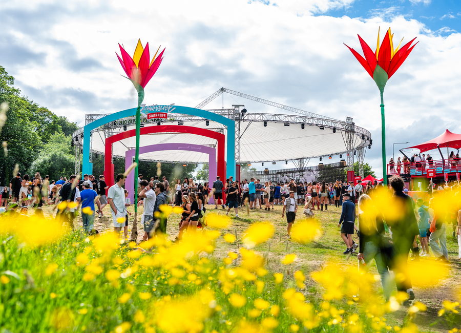 A festival main stage across a field of buttercups, giant inflatable flowers either side of a coloured arch and the crowd gathering in the sun